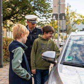 Zwei Schüler und ein Polizist stehen neben einem Auto am Straßenrand und reden mit einer Person im Auto.