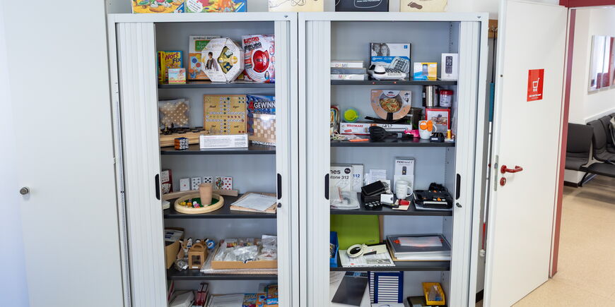 In einem Schrank mit Glastüren sind verschiedene Hilfsmittel und barrierefreie Spiele ausgestellt.
