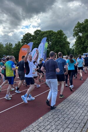 Auf einer Tartanbahn laufen Menschen in Sportkleidung, im Hintergrund sieht man Beachflags verschiedener Unternehmen. Ein Läufer dreht sich zur Kamera und hebt die Hände.