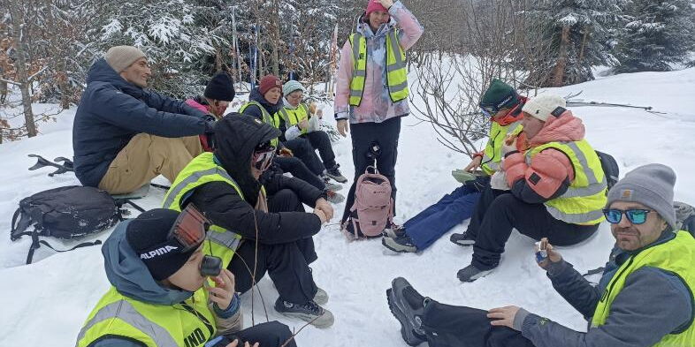 Vor einem winterlichen Wald sitzt die Gruppe aus Lehrkräften und Schüler*innen im Schnee und macht eine Verschnaufpause, viele in Skikleidung und Warnweste.
