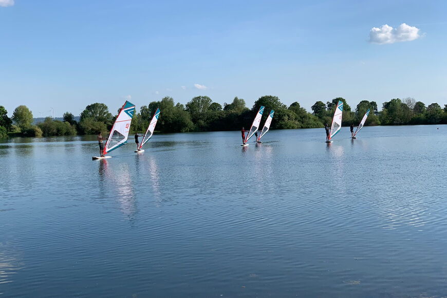 Viele Surfbretter von Ferne auf einem See, im Hintergrund Bäume und blauer Himmel.