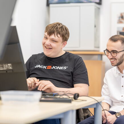 Zwei junge Männer sitzen gemeinsam an einem Pc und lächeln.