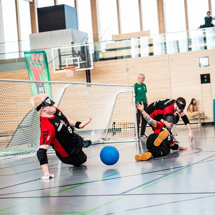 In einer Sporthalle werfen sich drei Goalballspieler vor ein Tor, um einen Ball abzuwehren.