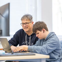 Eine Lehrkraft und ein Auszubildener sitzen an einem Laptop, der Azubi arbeitet mit einen Screenreader .