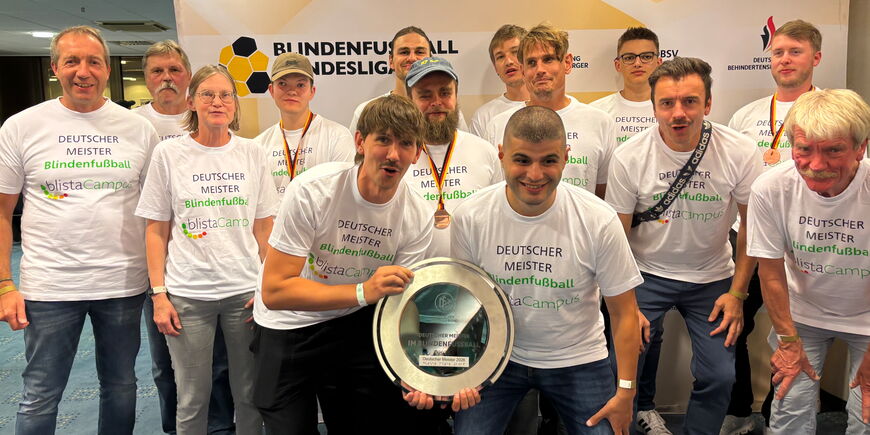Das Team der SF BG blista steht jubelnd gemeinsam mit dem blista-Vorstand und der Meisterplakette. Alle tragen weiße T-Shirts mit der Aufschrift Deutscher Meister Blindenfußball.