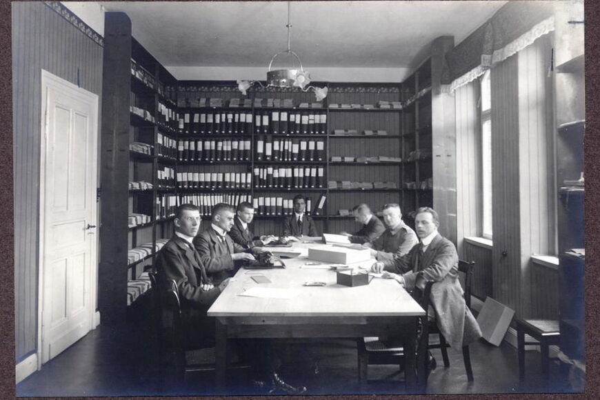 Auf dieser historischen Fotografie sieht man 7 Männer an einem Tisch sitzend, im Hintergrund sind hohe Regale mit Büchern zu sehen.