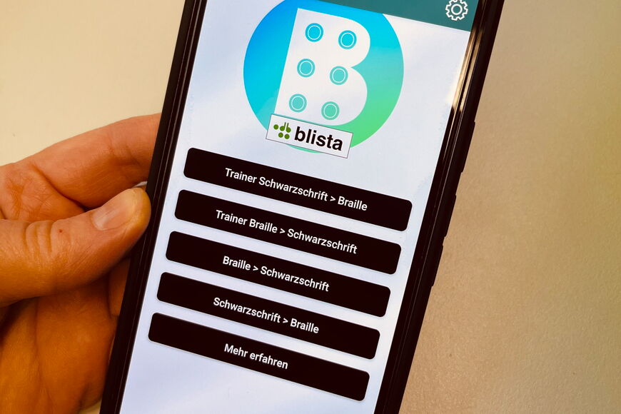 Eine Hand hält ein Smartphone, auf dem die Braille-Trainer App zu sehen ist.
