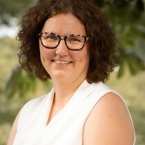 Portraitfoto  Melanie Grzimbke - sie hat braune Locken und trägt eine Brille. Sie trägt ein weißes Oberteil.