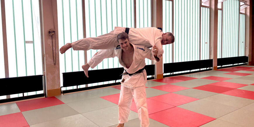 Zwei Judotrainer, die eine dynamische Judotechnik in einer Turnhalle auf Matten vorstellen.
