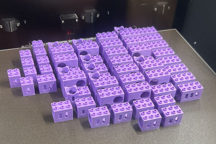 Auf der Druckplatte eines 3-D-Druckers liegen die selbstgedruckten fliederfarbenen Bauteile. Die Kunststoffsteine in doppelter Duplo-Höhe haben an verschiedenen Seiten runde Öffnungen, durch die die Kugel laufen kann.
