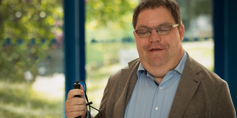 Thorsten Büchner trägt ein hellblaues Hemd, ein braunes Sakko und eine Brille. Er hat braunes Haar und einen Langstock in der Hand.