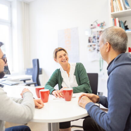In einem Büro sitzen drei Personen um einen Tisch, sie scheinen zu lachen.