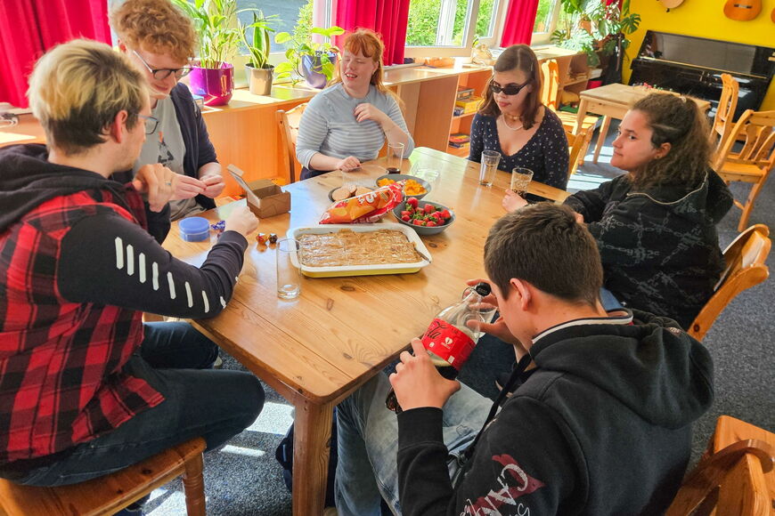 An einem Tisch in der Schüler*innen-Cafeteria sitzen sechs Personen, vor ihnen Snacks und verschiedene Würfel.