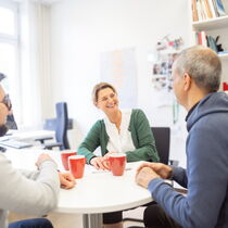In einem Büro sitzen drei Personen um einen Tisch, sie scheinen zu lachen.