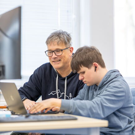 Eine Lehrkraft und ein Auszubildener sitzen an einem Laptop, der Azubi arbeitet mit einen Screenreader .