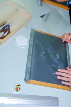 Eine Schülerin sitzt an einem Tisch, darauf ein Gallus-Brett. Darin ist eine Folie eingespannt. Auf der Folie sieht man Schrift, einen Kreis sowie ein Quadrat eingeprägt. Die Schülerin hält in der rechten Hand einen Griffel, mit der linken fährt sie über