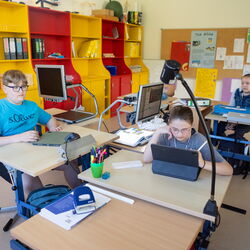 Drei Schüler*innen sitzen an ihren Arbeitsplätzen und arbeiten mit unterschiedlichen Medien.
