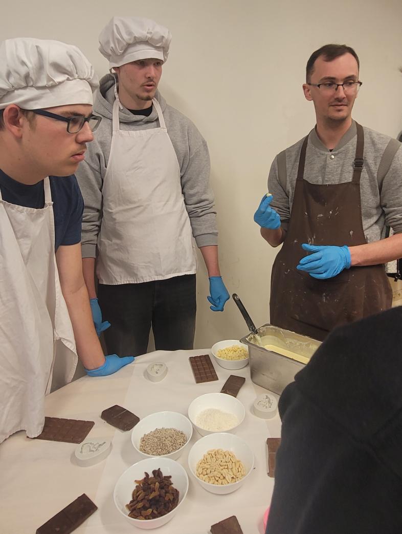 Wieder Schokolade, Werkzeuge und Zutaten auf Tischen, alle tragen blaue Handschuhe. Jannis und Hannes verfolgen in der Chocolaterie aufmerksam den Erläuterungen. 