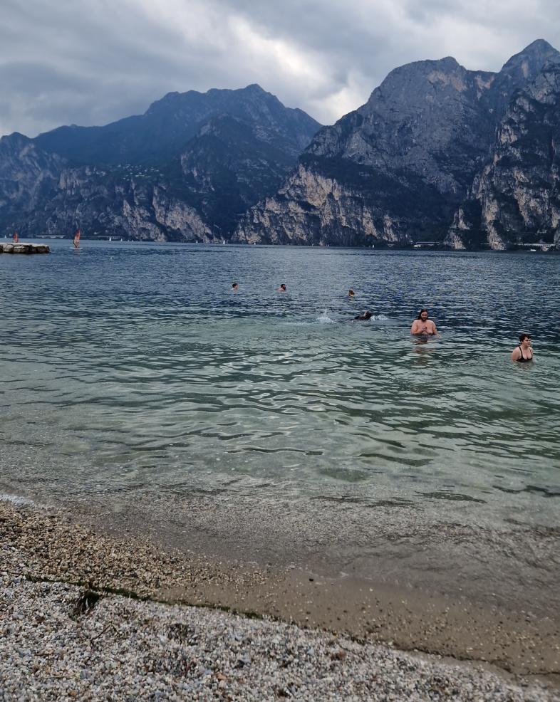 Ein paar Personen baden bei bewölktem Himmel im klaren Wasser des Gardasees. Im Vordergrund befindet sich ein Kiesstrand, im Hintergrund sieht man hohe Berge, die zum See hin ein Steilufer bilden