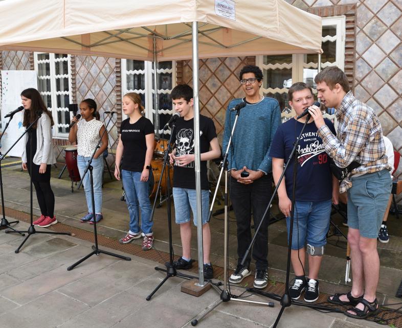 7 Schüler*innen der Schülerband der Carl-Stehl-Schule stehen im Innenhof des Museums unter einem Pavillon und singen in Mikrofone. 