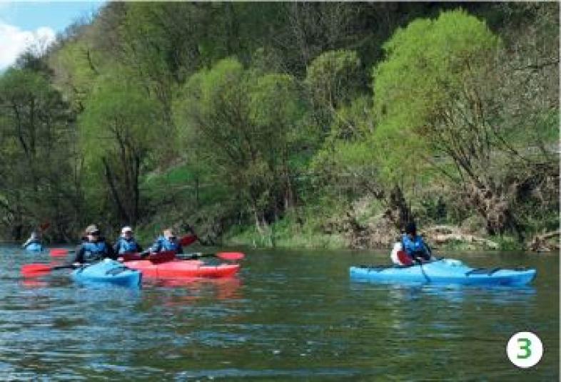 3.	Auf dem Fluss sieht man Menschen mit Kajaks entlangfahren, der Himmel ist blau mit wenigen weißen Wolken, an der Uferböschung frisch ausgetriebene Bäume. 