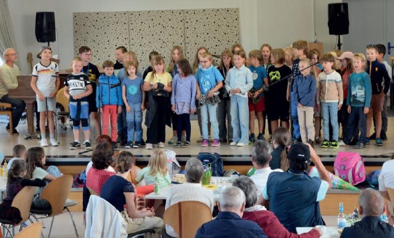 Eine Schulklasse steht auf der Bühne der Aula und begrüßt musikalisch die neuen Mitschüler*innen, die mit ihren Familien im Publikum zusehen.