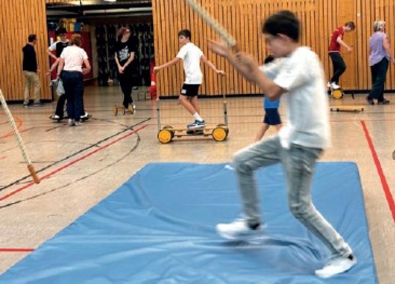 Jede Menge sportliche Aktivität in der blista-Sporthalle. Im Vordergrund spielt ein Kind mit einem von der Decke hängenden Seil auf einer Matte, im Hintergrund fahren Kinder auf Pedalos.