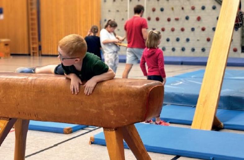 Mehrere Kinder spielen in der Turnhalle, im Vordergrund klettert ein Kind auf einen Turnbock.