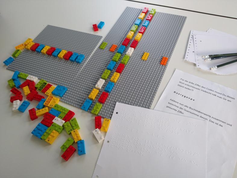 Auf einem Tisch liegen Zettel in Schwarz- und Punktschrift sowie bunte Lego Braille-Bricks und Bleistifte. 