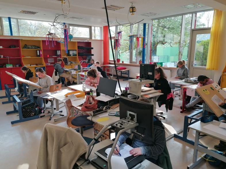 1.	In einem Klassenraum sitzen Schüler*innen vor Tischen, manche mit Bildschirmlesegeräten und arbeiten konzentriert an den kniffligen Aufgaben des Kanguru-Wettbewerbs.