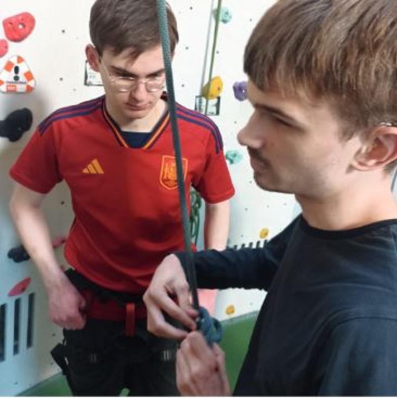 Zwei Schüler in Sportkleidung stehen vor einer Wand mit Klettergriffen. Einer hat ein Kletterseil in der Hand, während sein Kletterpartner ihm beim ertasten des Knotens zusieht.