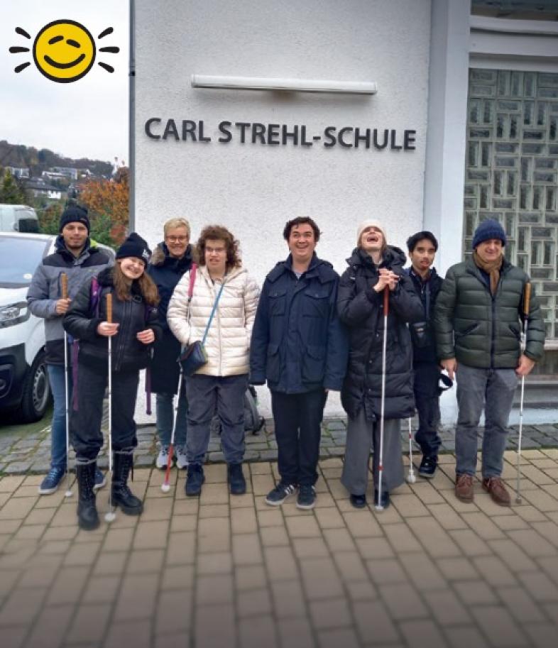 Acht fröhliche Menschen mit und ohne Langstöcke stehen winterlich gekleidet vor dem Schild Carl-Strehl-Schule.