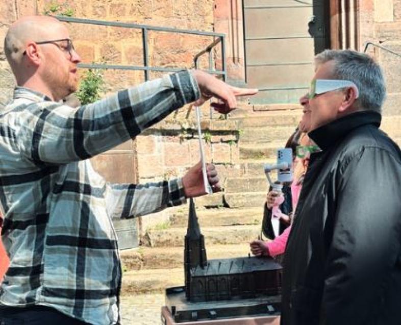 Low-Vision-Experte Martin Brehmer zeigt Oberbürgermeister Spies, der eine Simulationsbrille trägt, eine Sehtafel.