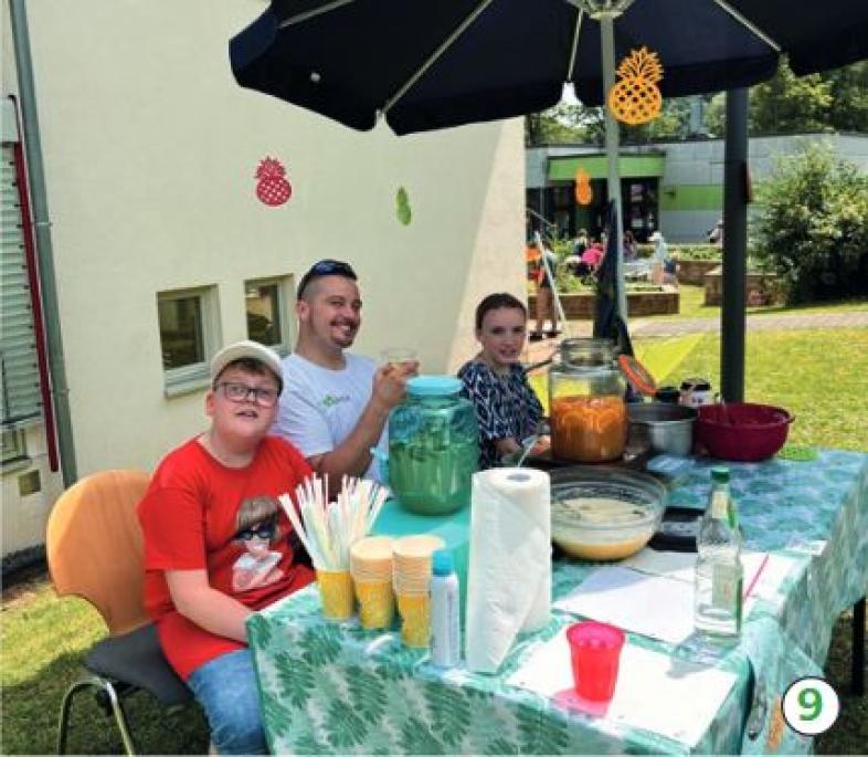 9.	Beim Cocktailstand der sechsten Klasse sitzen zwei Schüler*innen mit ihrem Lehrer unter einem Sonnenschirm vor großen Cocktailgefäßen.